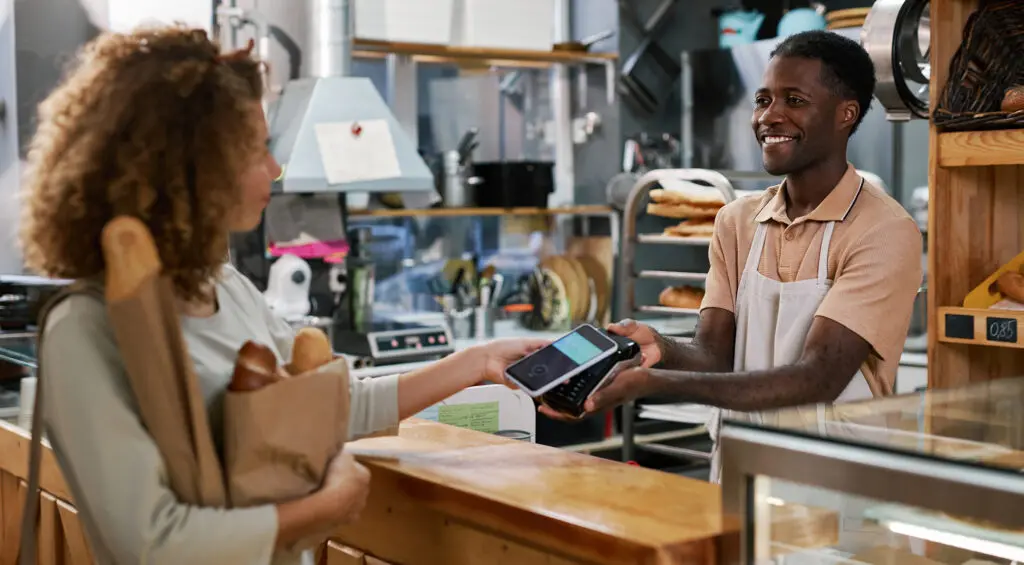 A woman holding a bag of bread at a bakery pays at the POS terminal using her smartphone.